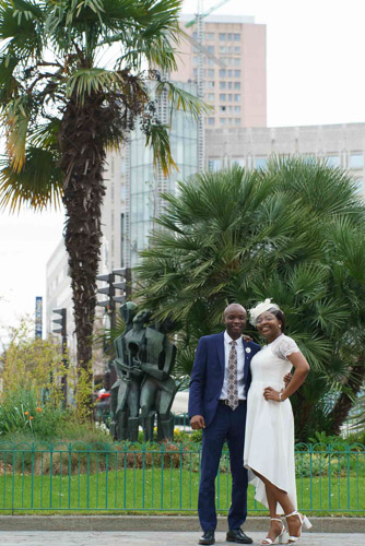 Photo mariage civil Paris 13e – Mariés debout côte à côte, souriant devant un palmier sur le parvis de la mairie