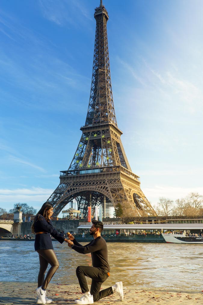 Paris proposal photographer surprise, joy on fiancée's face, fiancé placing ring on her finger, Eiffel Tower, Seine riverside, Port Debilly, Paris, winter afternoon, blue sky, golden sunlight